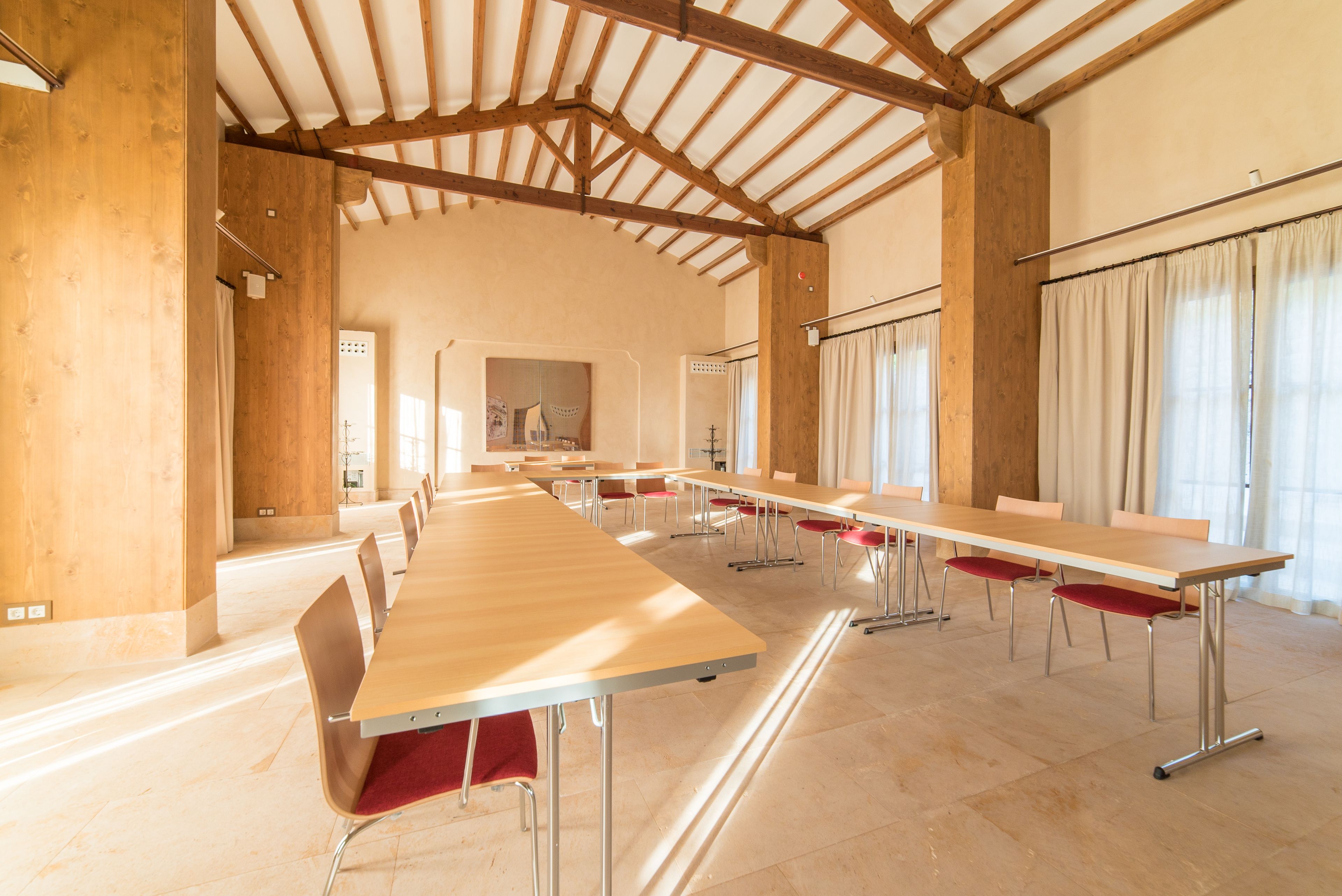A large conference room with long tables and chairs, wooden beams on the ceiling, and large windows with curtains.
