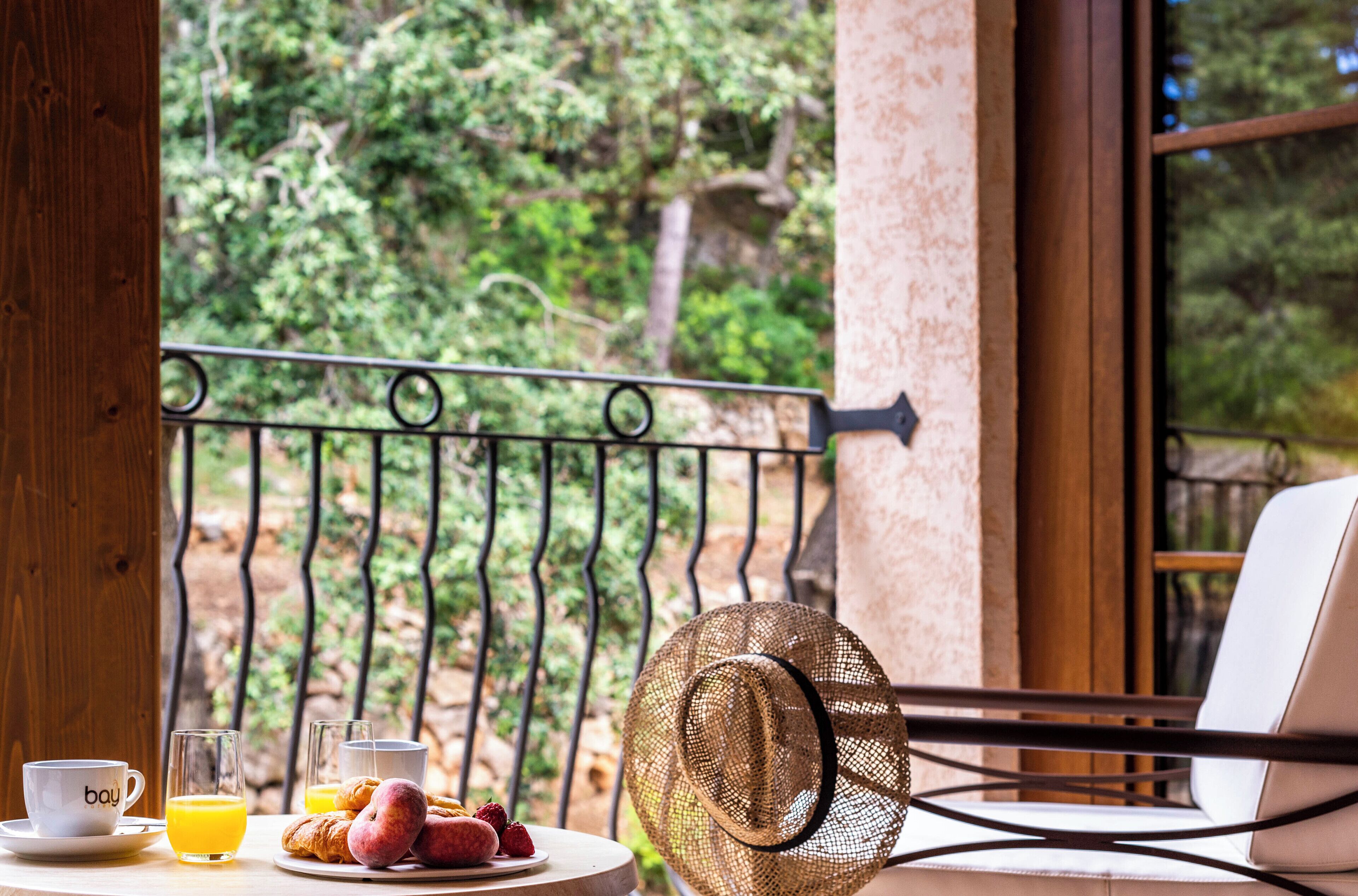 Una mesa de desayuno con cruasanes, un sombrero y una computadora portátil en un balcón con vista a árboles.