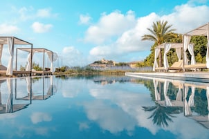 Una piscina cristalina que refleja el cielo y palmeras, rodeada de zonas de descanso a la sombra.