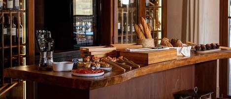 A bar area with a wooden counter, stools, wine bottles, glasses, and pastries.