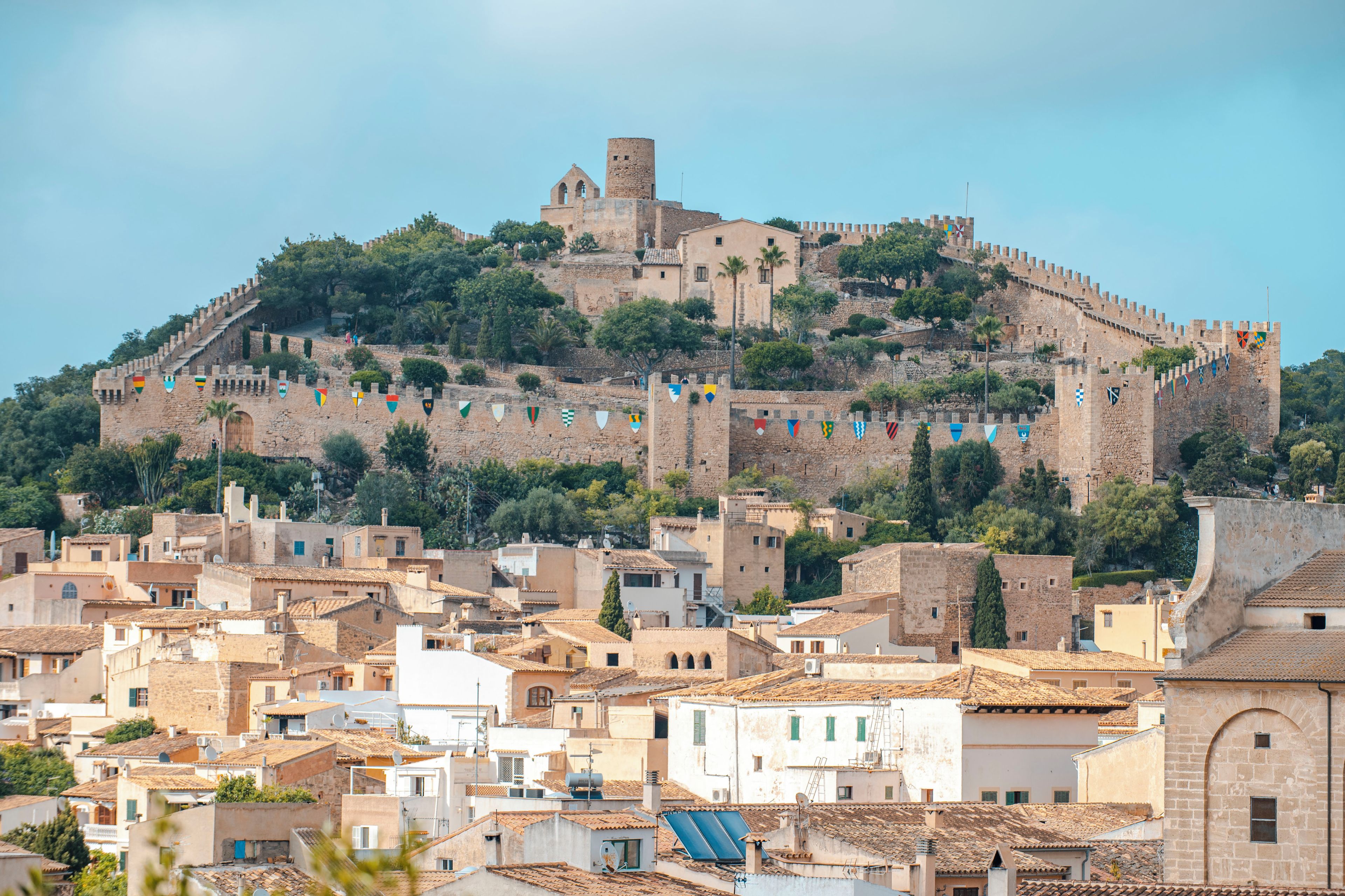 Um castelo histórico numa colina, com uma muralha fortificada e bandeiras, com vista para uma vila de arquitetura tradicional.