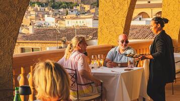 An outdoor dining area with tables and chairs, a waiter, and a view of a historic town in the background.