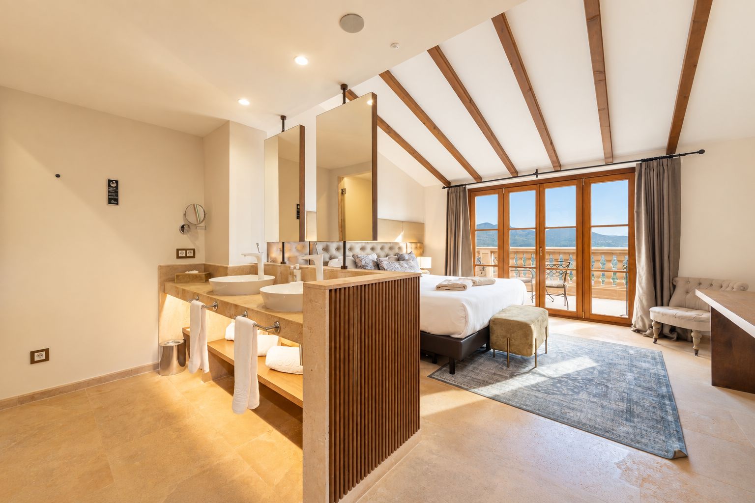 A modern hotel room with a large bed, a sitting area, a bathroom with two sinks, and a balcony with a view.