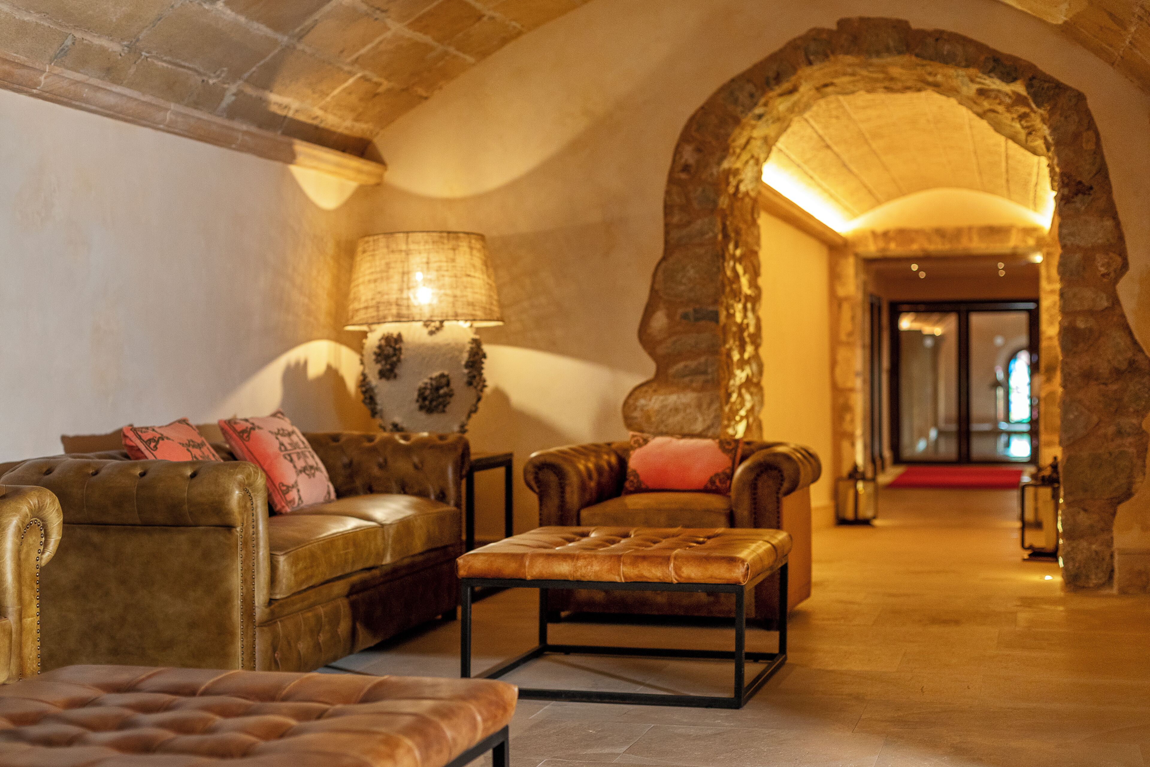 A cozy lounge area with leather sofas and a tufted ottoman, a stone archway, and a lamp with a patterned lampshade.
