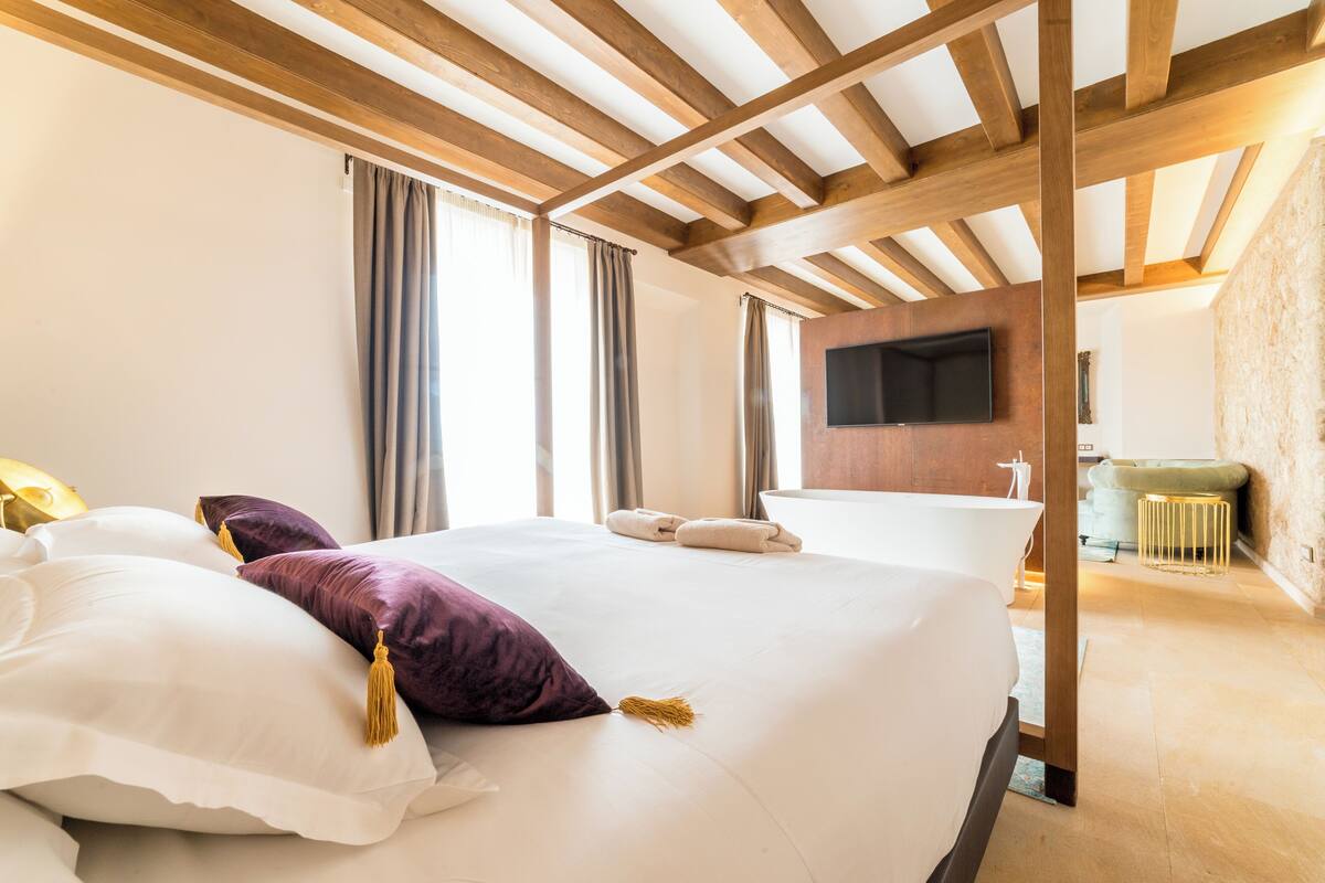 Quarto de hotel com cama grande, vigas de madeira no teto, televisão de tela plana e banheiro visível ao fundo.