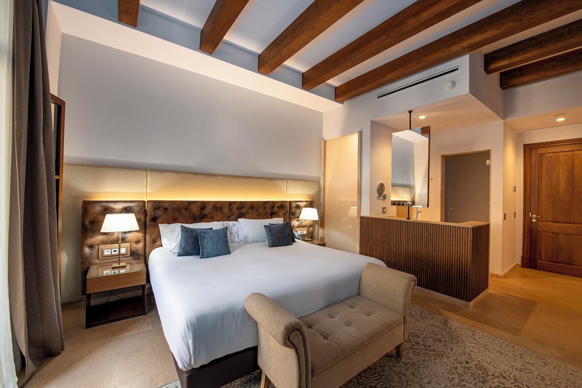 Quarto de hotel moderno com uma cama grande, luminárias de cabeceira, cabeceira de madeira, um sofá, um espelho e um balcão de recepção de madeira.