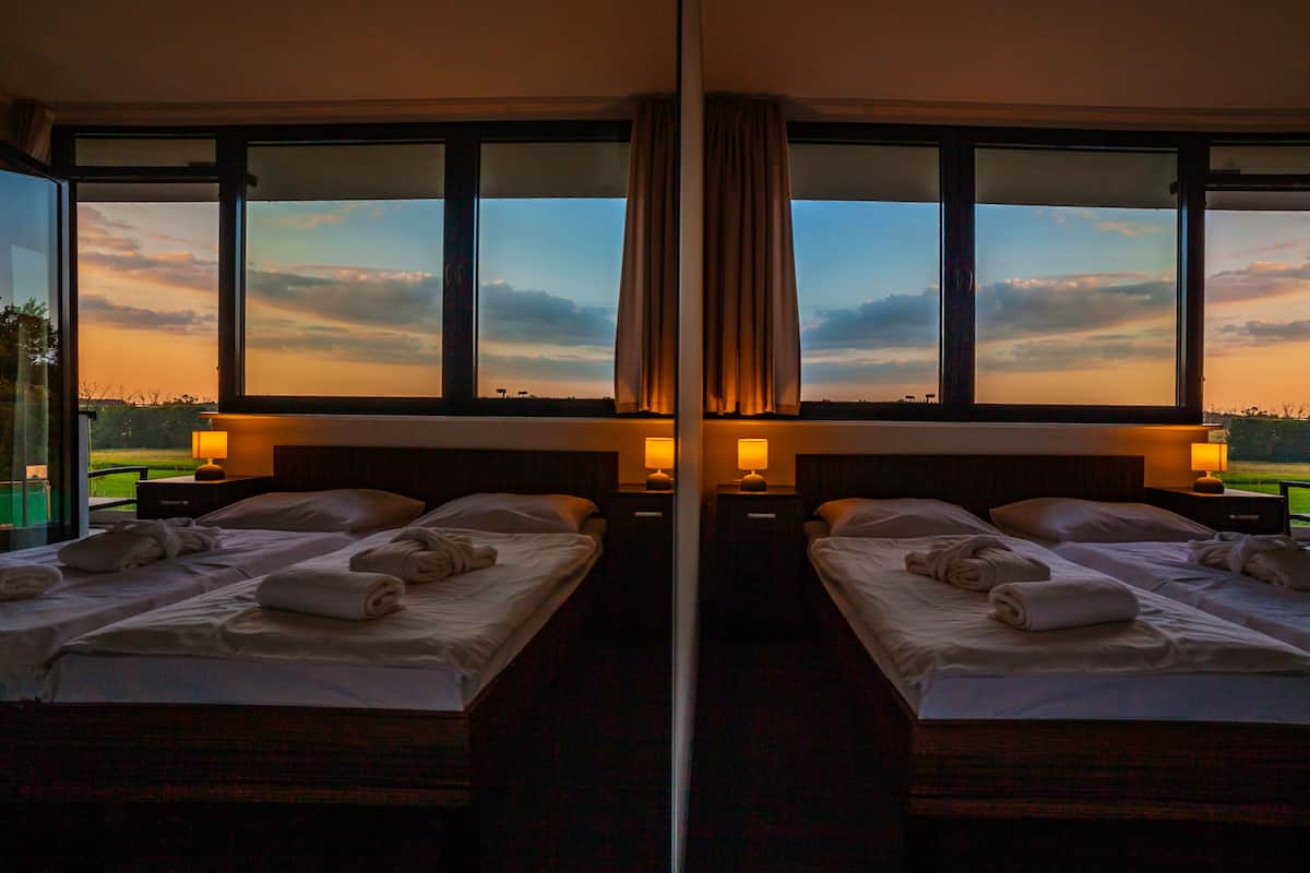 Ein Hotelzimmer mit Spiegel, zwei Betten, jeweils mit einem Kissen, einem Nachttisch mit Lampe und Blick auf einen Sonnenuntergang durch große Fenster.