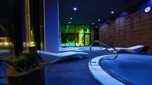 Una piscina interior moderna con paredes de vidrio transparente, una pared de ladrillo en un lateral y un área de descanso con tumbonas blancas.