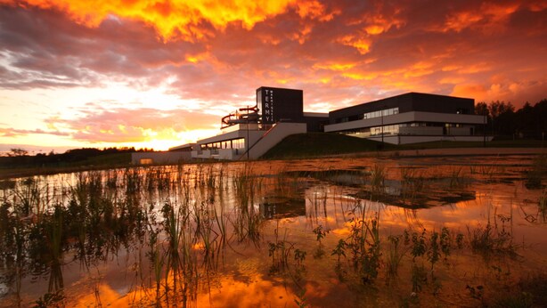 Un edificio moderno de techo plano y forma rectangular, ubicado junto a una masa de agua que refleja el cielo.