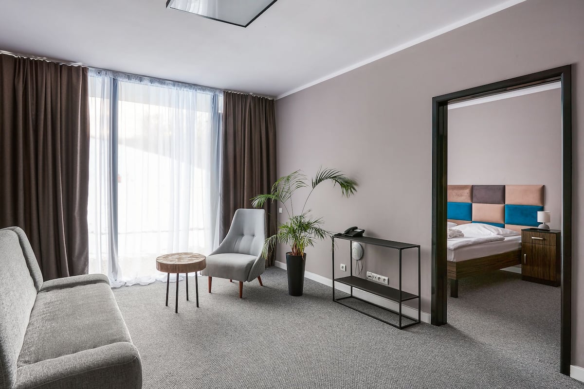Ein Hotelzimmer mit grauem Sofa, einem kleinen runden Tisch, einem Blumentopf, einem Nachttisch mit Lampe und einem Bett mit blau-beiger Kopfteil.