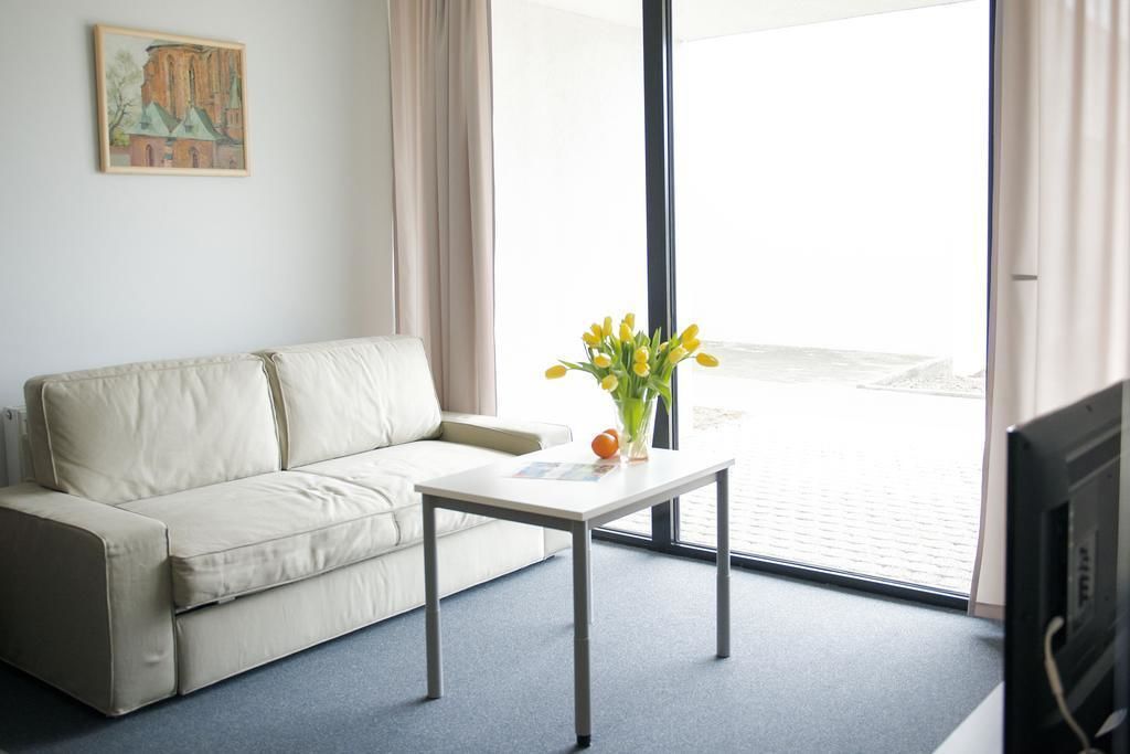 A room with a beige sofa, a small white table with a vase of yellow flowers, and a large window with a view of a building.