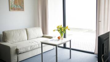 A room with a beige sofa, a small white table with a vase of yellow flowers, and a large window with a view of a building.