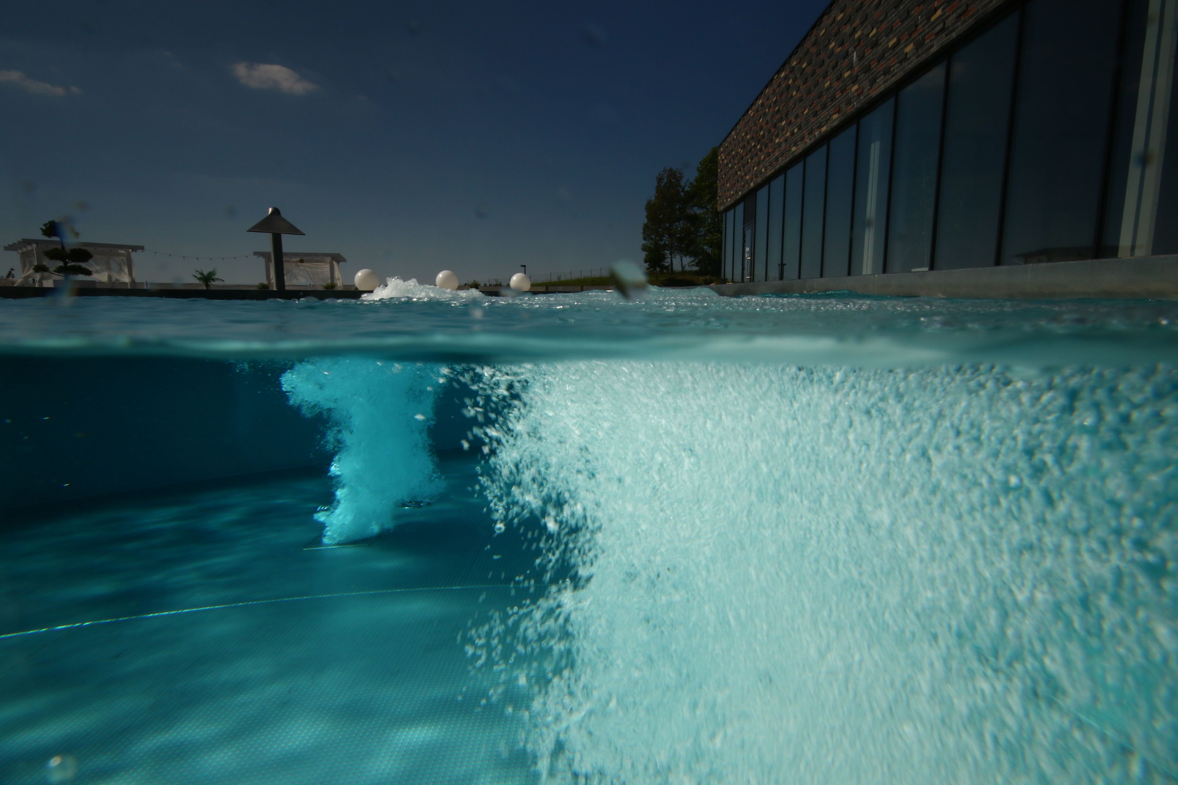 Una piscina con agua cristalina, una ola y un edificio con grandes ventanales al fondo.
