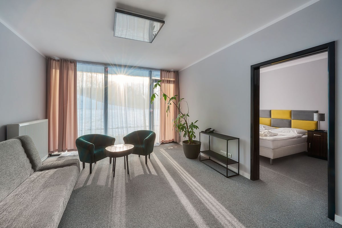 Ein modernes Hotelzimmer mit einer Couch, zwei Sesseln, einem kleinen Tisch, einer Topfpflanze, einem Bett mit gelbem Kopfteil und einem Nachttisch.
