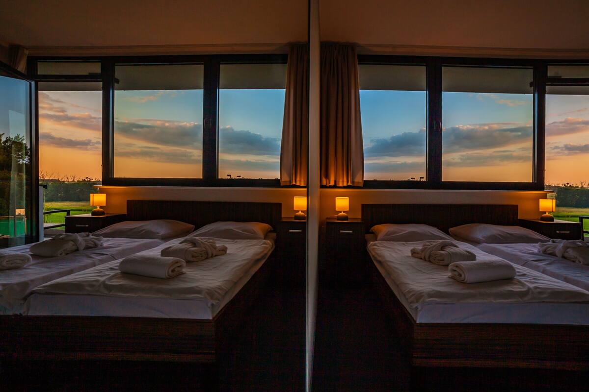 Ein Hotelzimmer mit Spiegel, ausgestattet mit zwei Betten, jeweils mit einem Kopfkissen, einem aufgerollten Handtuch und einem Nachttisch mit einer Lampe.
