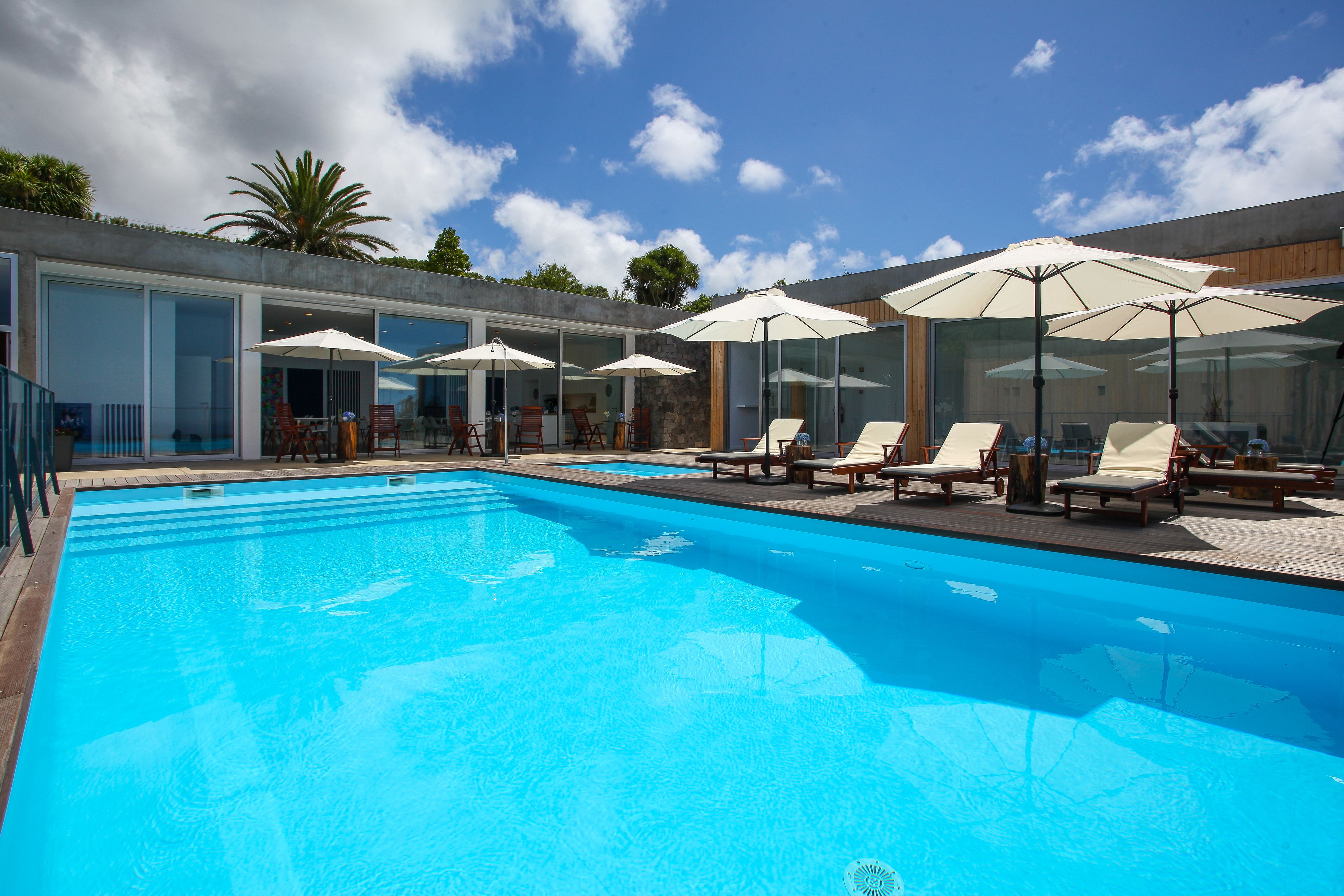 Piscina con sillas de descanso y sombrillas, rodeada de paredes de vidrio y una terraza de madera.