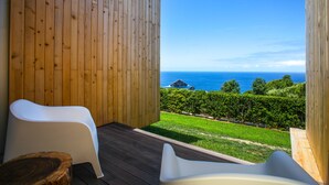 A wooden deck with two white chairs, a small table, and a view of the ocean and greenery.