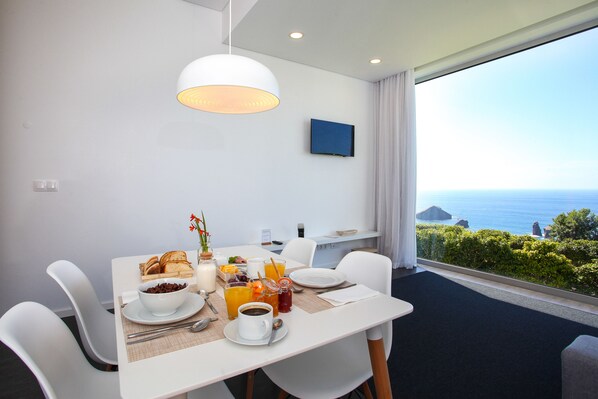 A dining area with a table set for breakfast, including bread, coffee, and fruit. A large window offers a view of the sea and greenery.