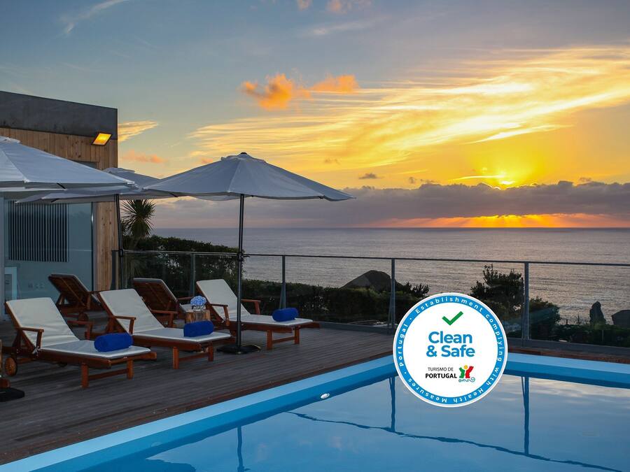 Zona junto a la piscina con sillones reclinables y sombrillas, con vista al mar al atardecer.