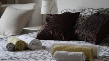 Un letto rifatto con cura, lenzuola a fantasia, asciugamani arrotolati nei colori bianco, giallo e grigio, e una lampada da comodino.