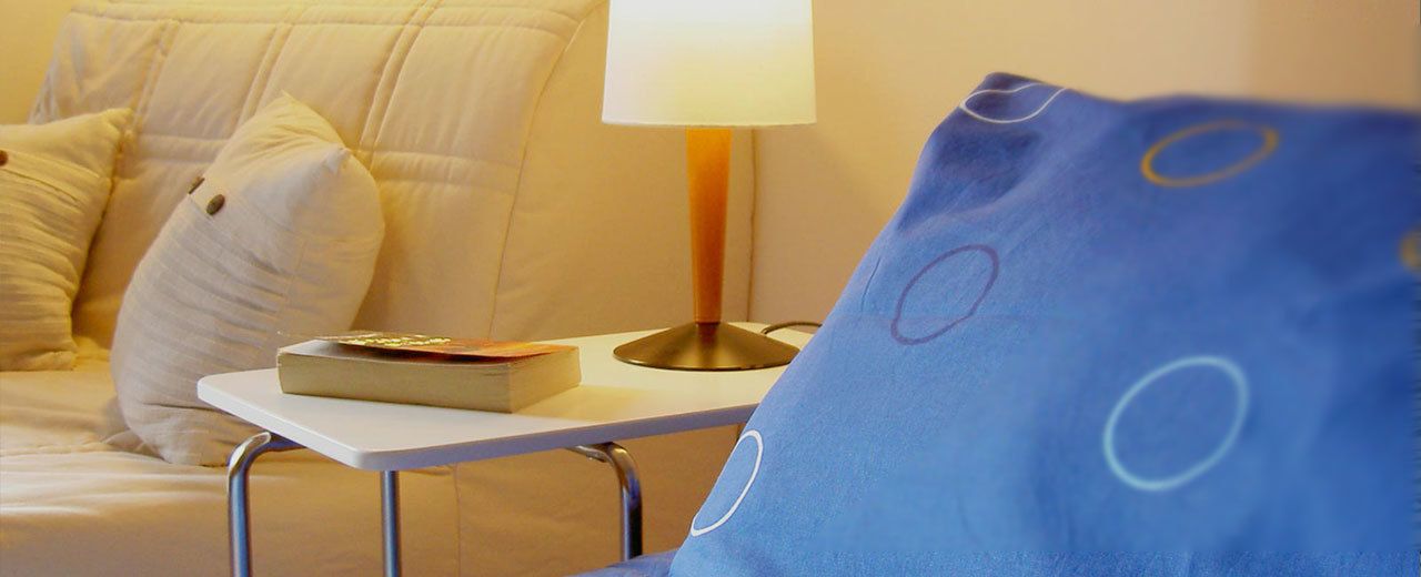 Ein ordentlich bezogenes Bett mit einem weißen Kopfkissen, ein Nachttisch mit Buch und Lampe sowie ein blaues Kissen mit kreisförmigem Muster.