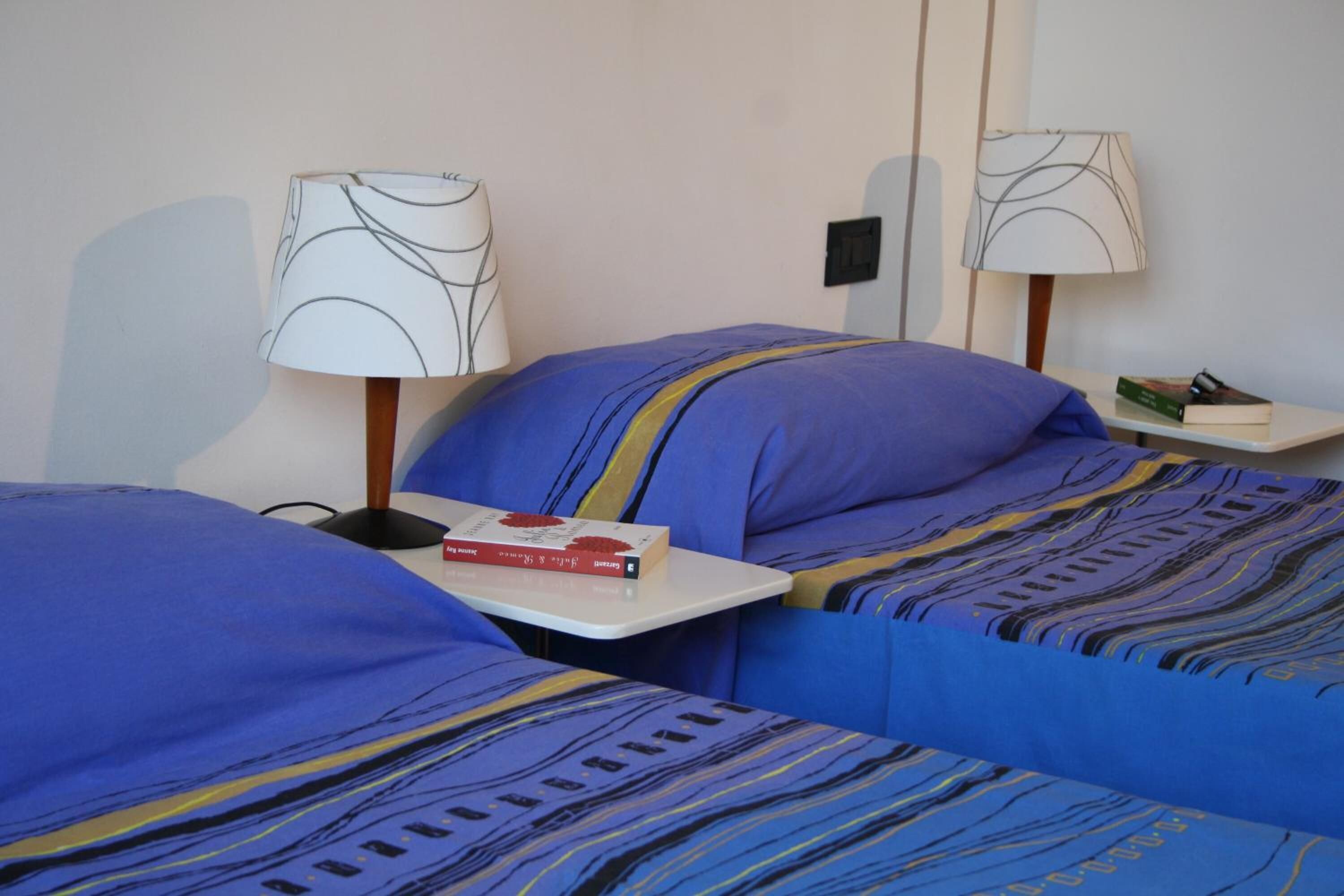 A hotel room with two beds, each with a blue bedspread and a matching pillow. There are two bedside tables, each with a lamp and a book.