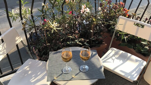 Una mesa con dos copas de vino sobre un tapete, rodeada de sillas y una planta en maceta con flores.