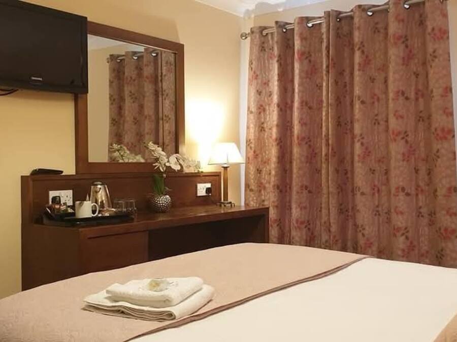 Une chambre d’hôtel avec un lit, une table de chevet avec une lampe, un miroir, une télévision et des rideaux à motifs floraux.