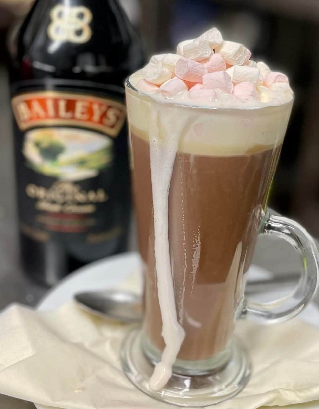A tall glass mug with a layered drink topped with marshmallows, a bottle labeled "Bailey's" in the background.