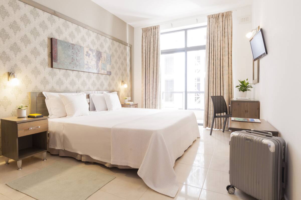 A hotel room with a large bed, bedside tables, a desk, a chair, a TV, and a window with curtains.