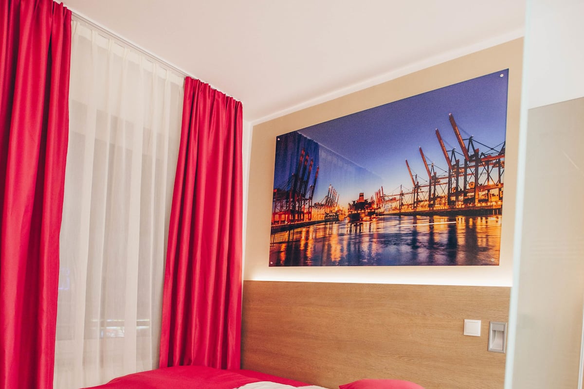 A hotel room with a bed, red curtains, a large window, and a cityscape painting on the wall.