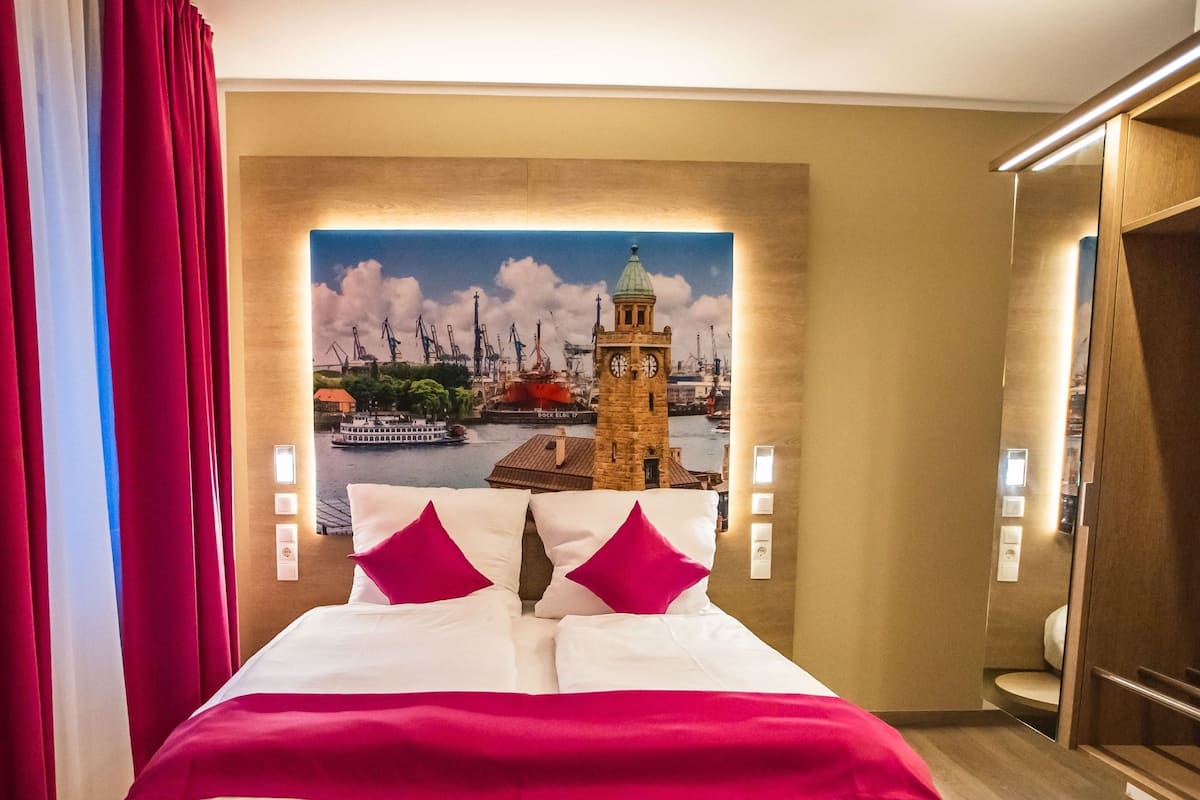A hotel room with a bed, red curtains, a large window with a view of a clock tower, and a wooden wardrobe.
