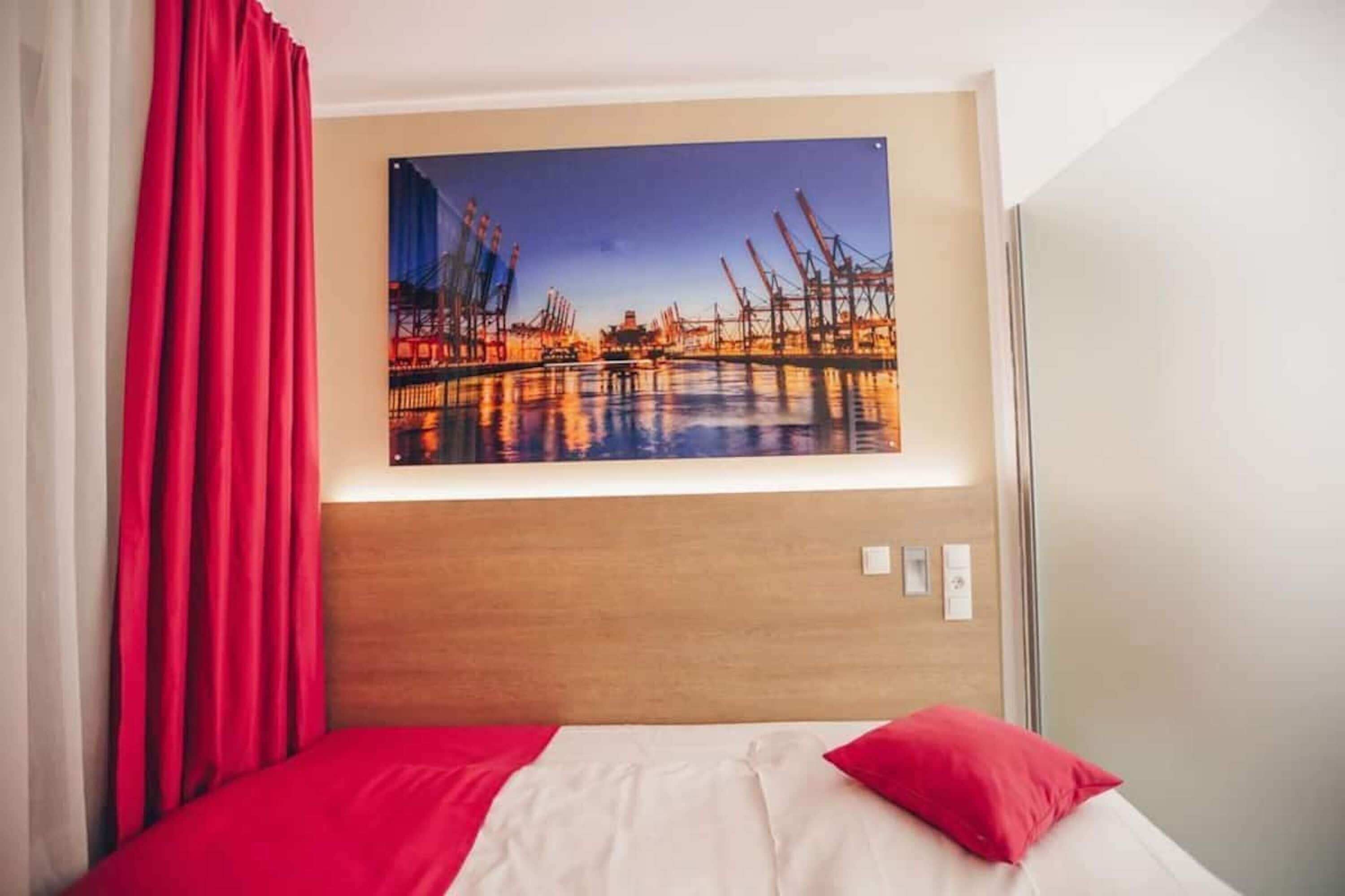 Een hotelkamer met een bed, rode gordijnen, een houten hoofdeinde en een groot schilderij van een waterlandschap.
