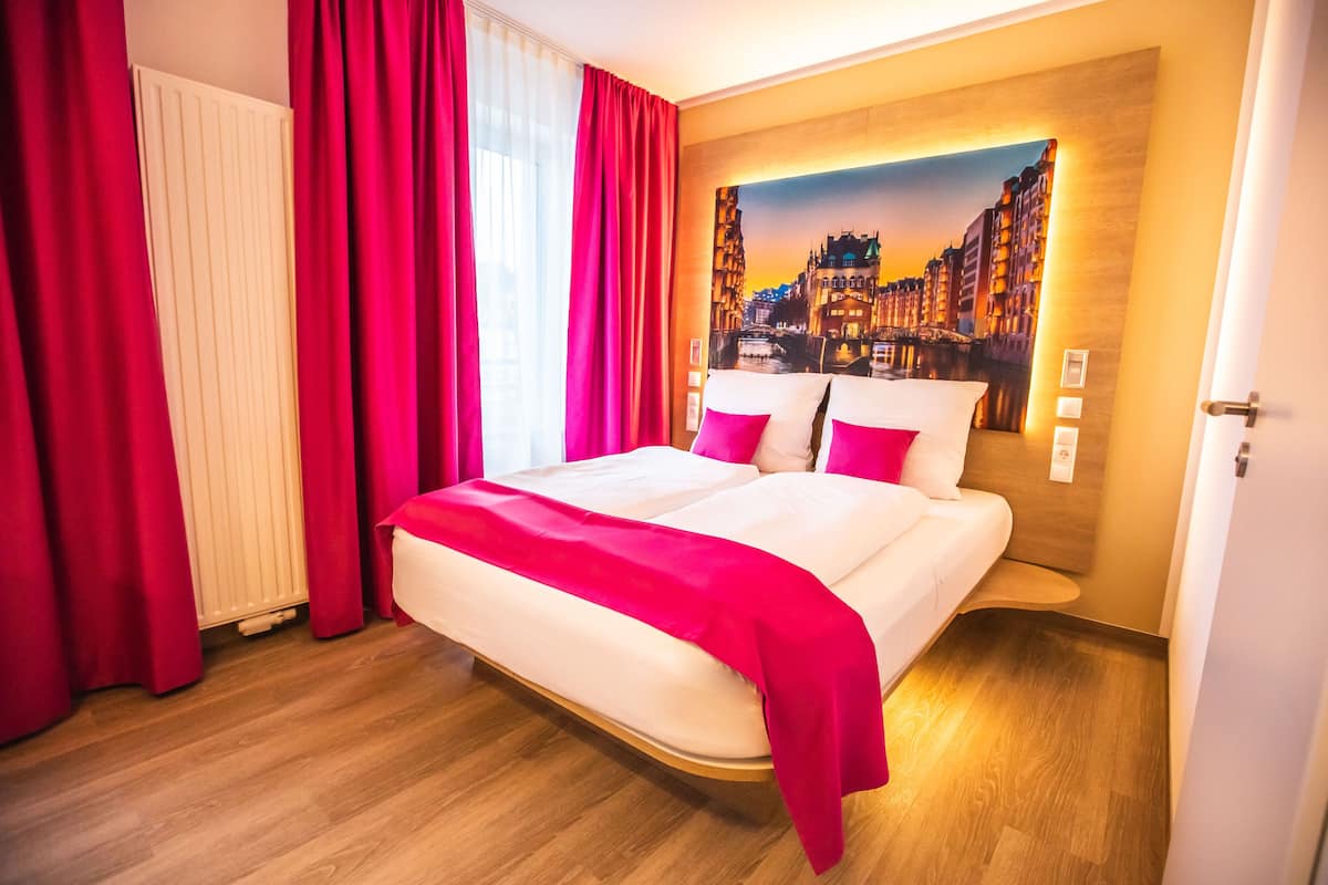 A hotel room with a large bed, red curtains, a cityscape mural on the wall, and wooden flooring.