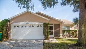 A single-story house with a white garage door, a covered driveway, and a small front yard with a tree and shrubbery. - Ly58134 - Indian Ridge - 4 Bed 2 Baths Villa (Kissimmee)