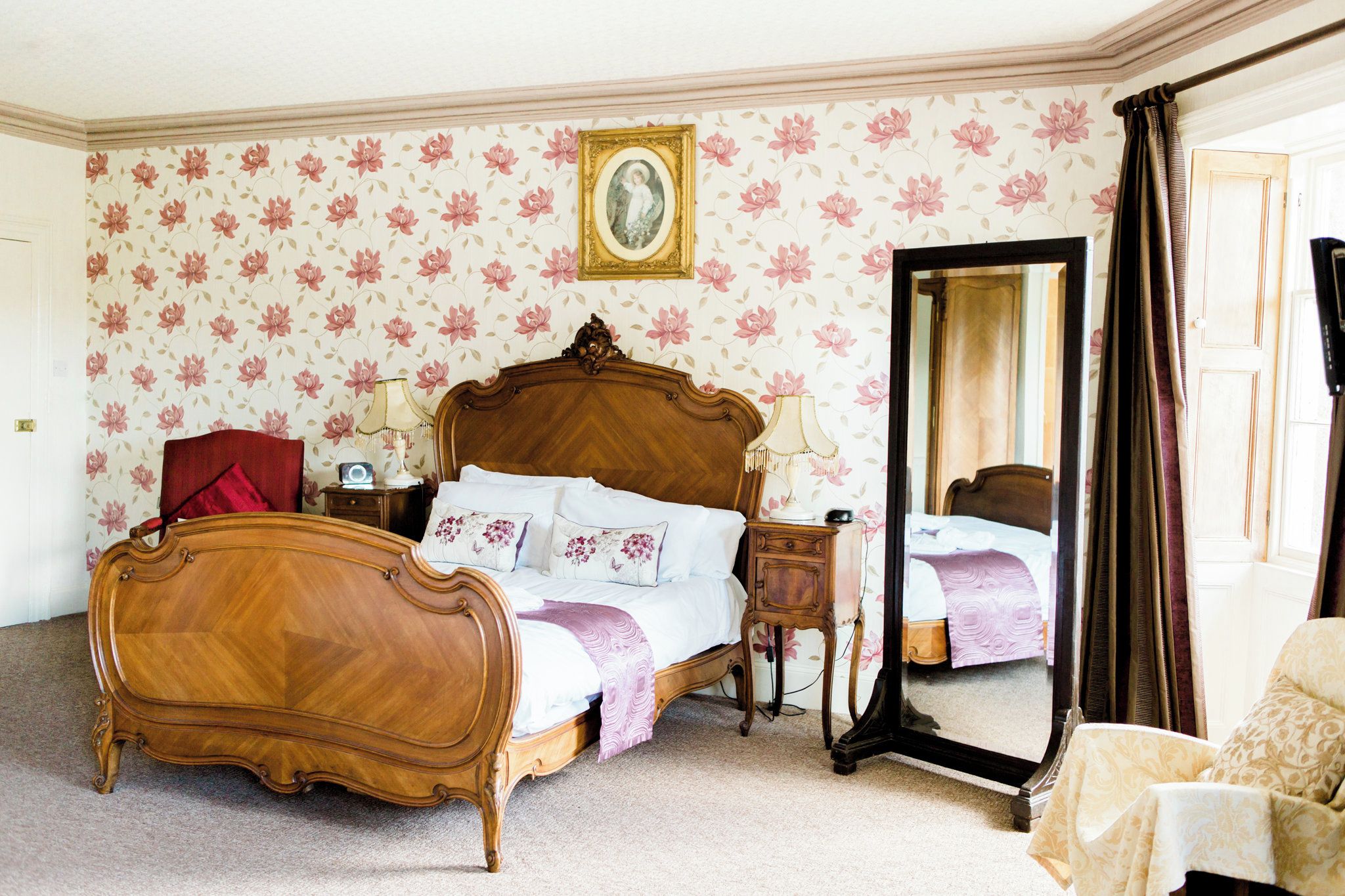 A bedroom with a wooden bed, a nightstand, a mirror, and floral wallpaper.