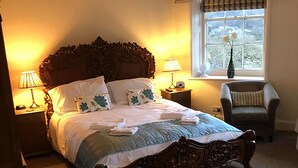 Een opgemaakt bed met een houten hoofdeinde, twee bedlampjes, een raam met uitzicht en een stoel in een kamer met beige wanden.