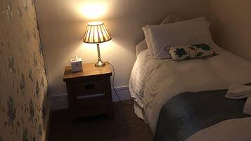 A small, dimly lit bedroom with a single bed, a bedside table with a lamp, and floral wallpaper.