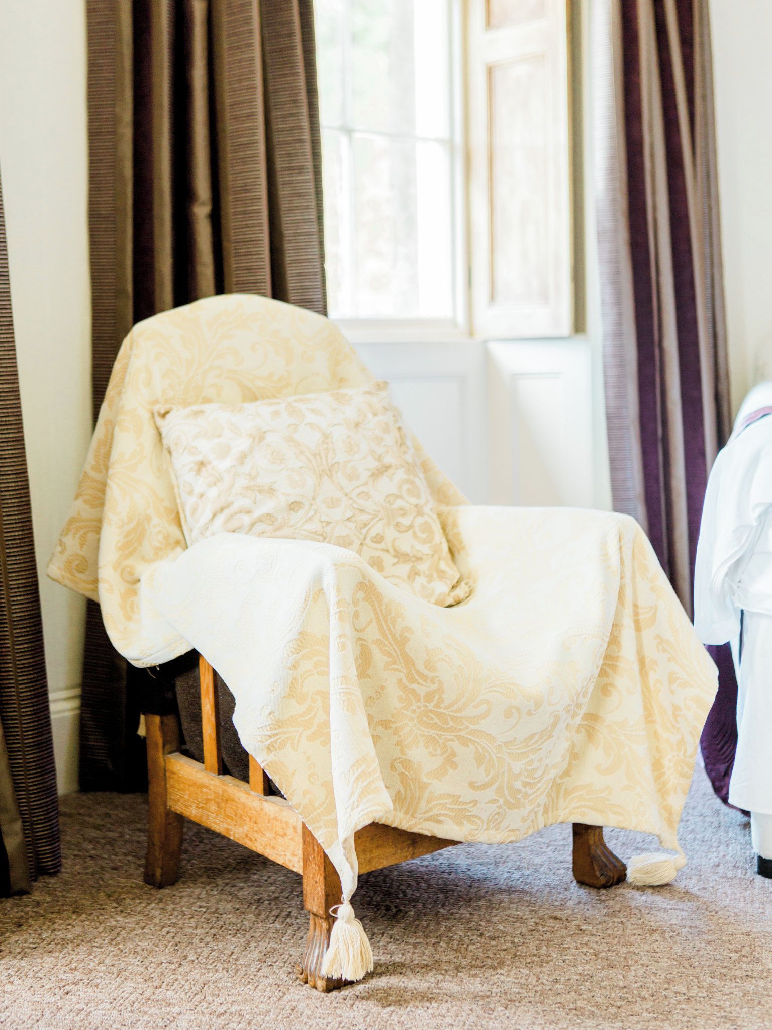 A room with a single wooden chair covered in a patterned blanket, a window with curtains, and a carpeted floor.