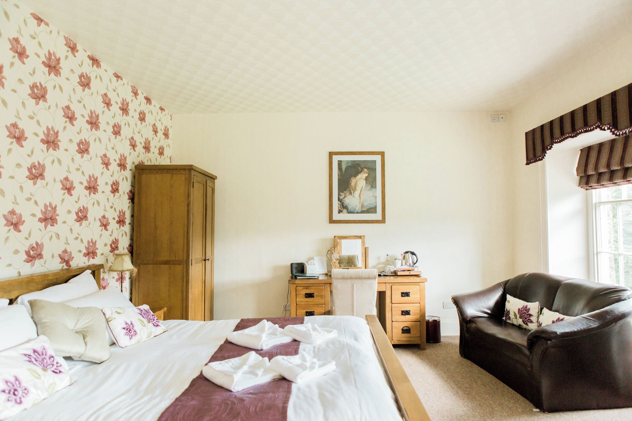 A bedroom with a bed, a wooden wardrobe, a desk with a chair, a painting on the wall, and a window with curtains.