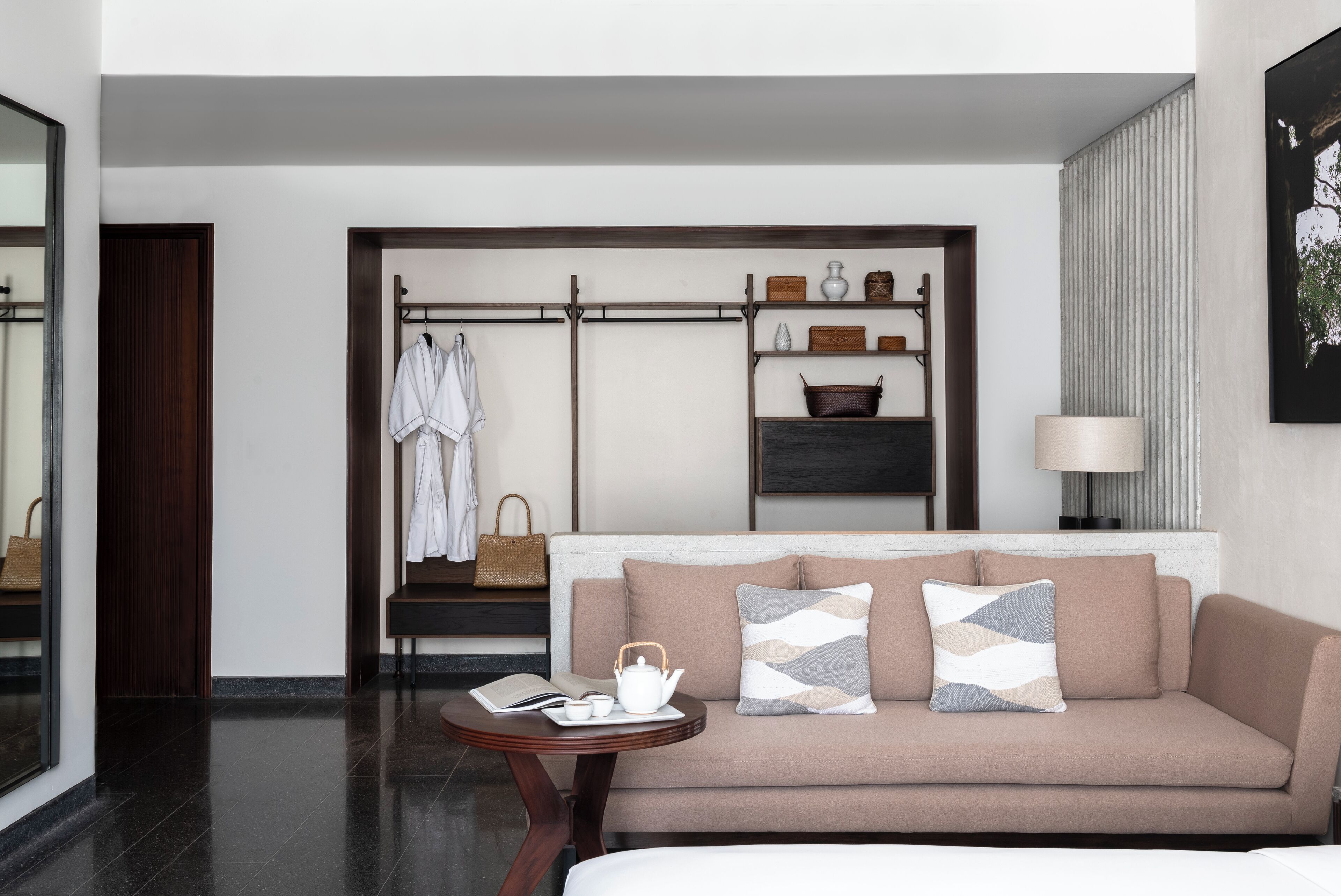 A modern hotel room with a beige sofa, a small round table with a teapot and book, a coat rack with a robe, and a shelf with decorative items.