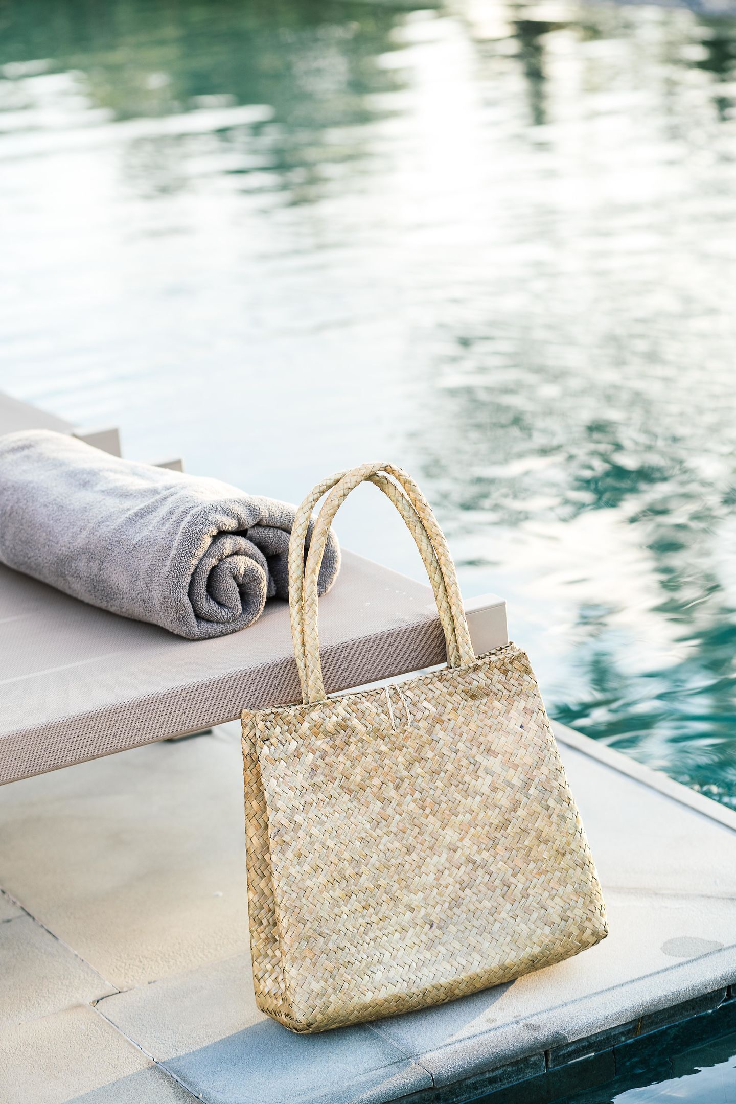 Un bolso tejido sobre una silla de salón junto a una piscina, con una toalla enrollada al lado.
