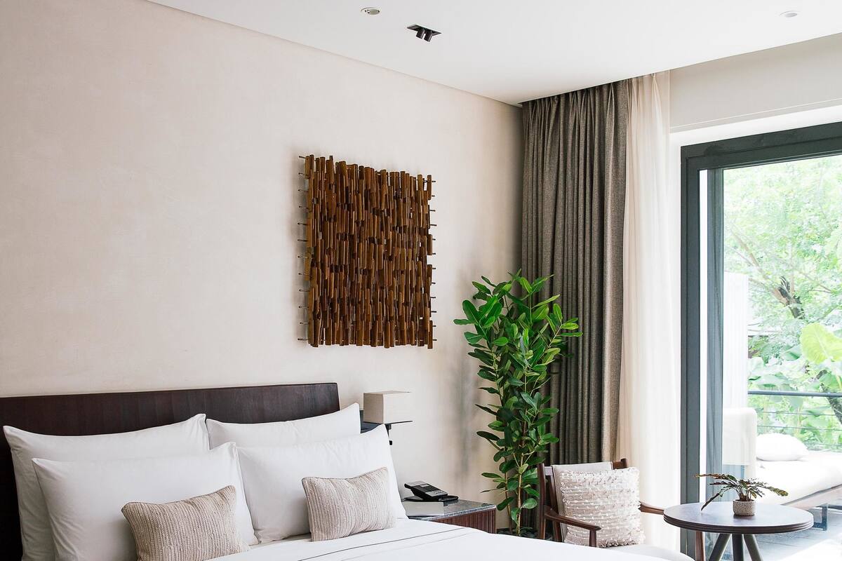 A modern hotel bedroom with a large bed, a bedside table, a chair, a potted plant, and a view of greenery outside.