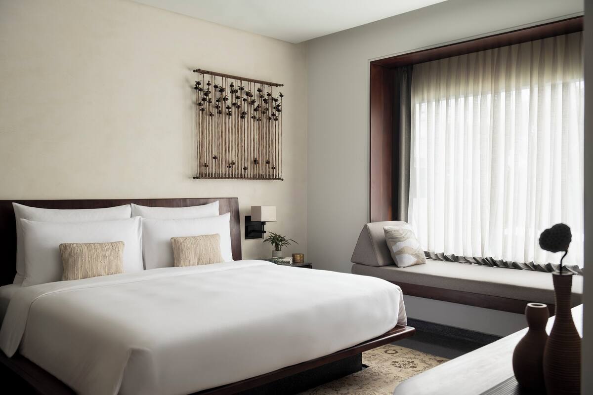 A hotel room with a large bed, a bedside table with a lamp, a window with blinds, and a decorative wall hanging.
