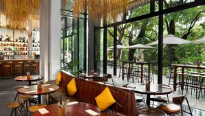 An indoor dining area with wooden tables and benches, large windows offering a view of trees, and a bar with shelves of bottles. - Treeline Urban Resort (Siem Reap)