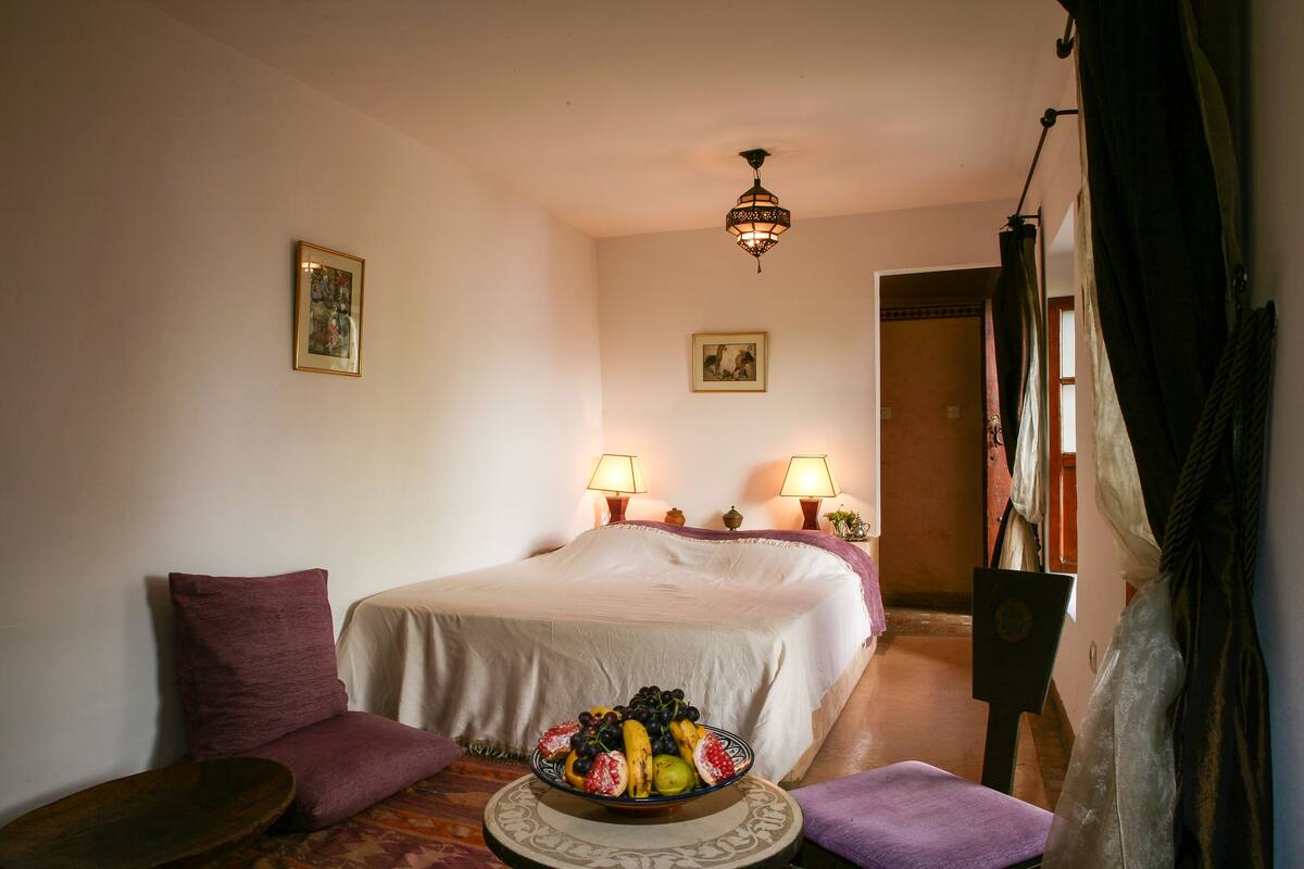 Habitación de hotel con una cama, dos lámparas de noche, un frutero sobre la cama, una mesita redonda y una silla.