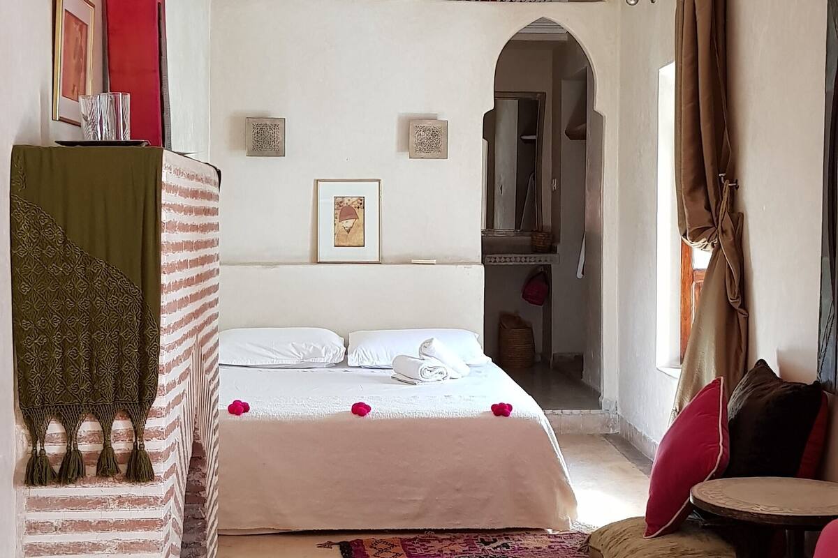 Un dormitorio con cama, una mesita, una silla, una lámpara colgante y un tapete estampado.