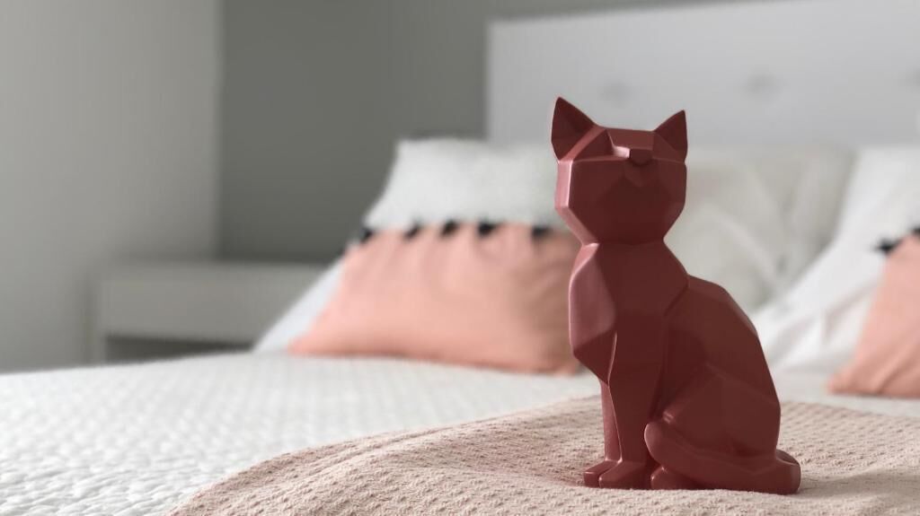 A 3D cat figurine on a bed with a white and pink pillow arrangement.