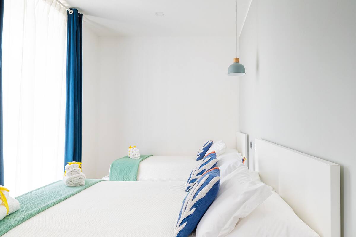A modern hotel room with a large bed, white linens, a blue and white pillow, a green towel, and a window with blue curtains.