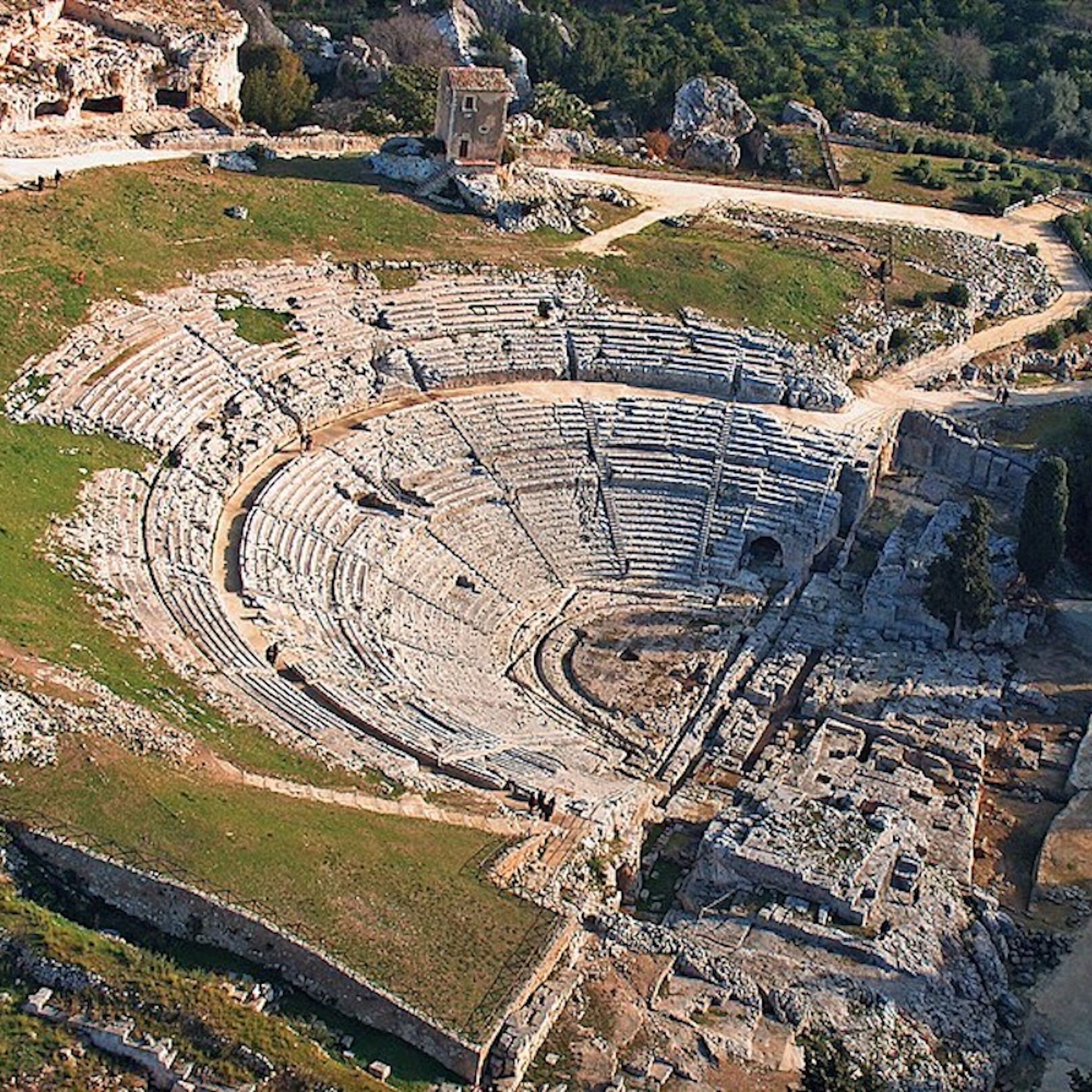 An ancient amphitheater with tiered seating and a central stage, surrounded by ruins and greenery.